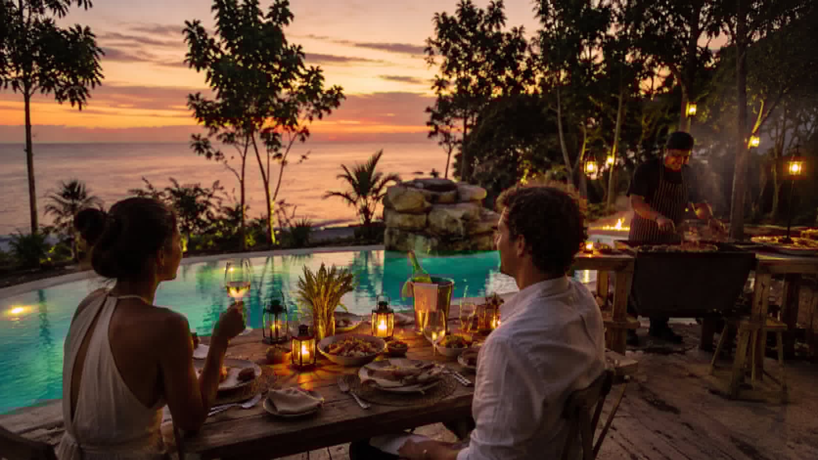 Two guests dining at a long table beside the pool at sunset.